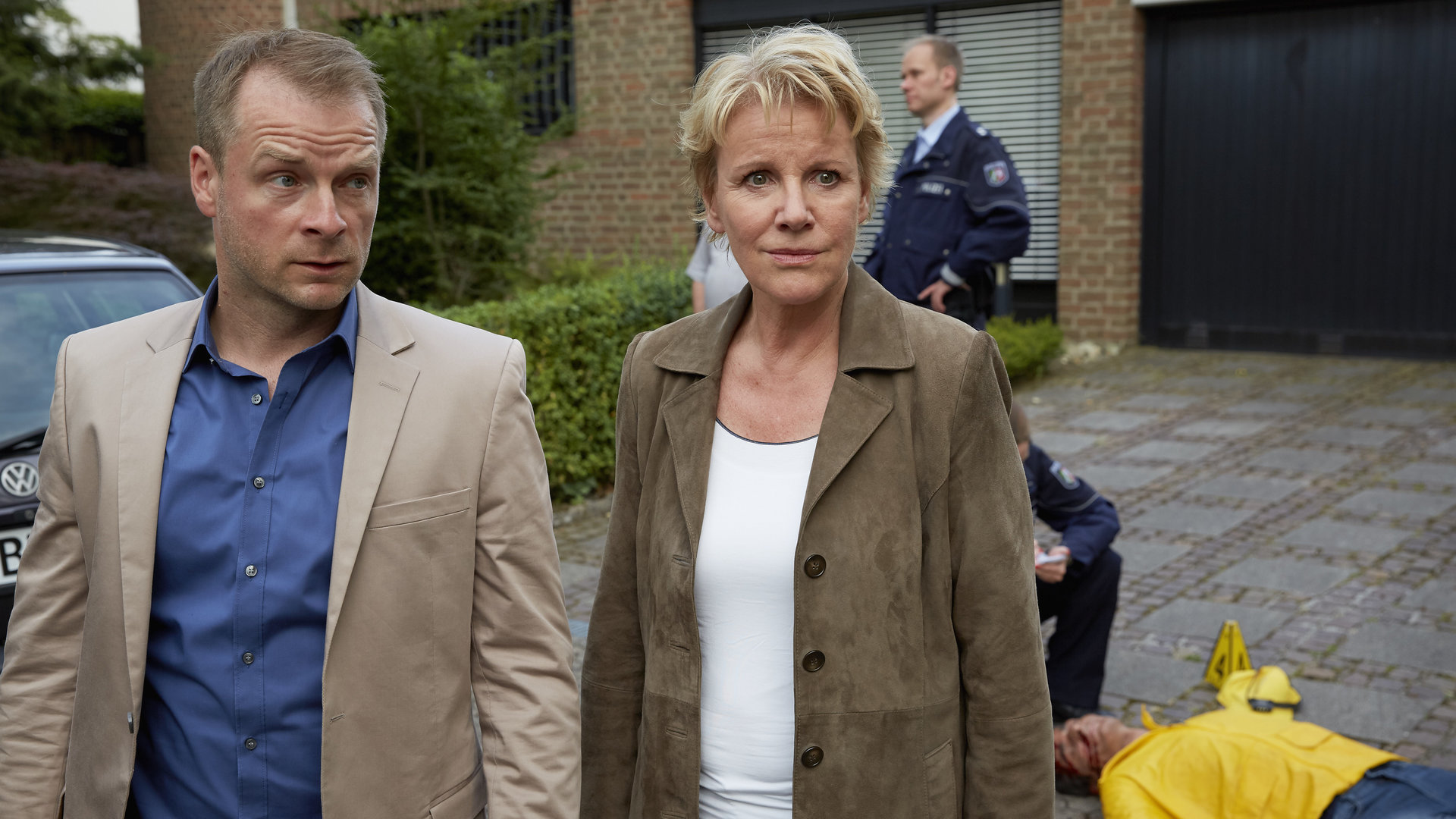 "Marie Brand und das ewige Wettrennen": Marie Brand (Mariele Millowitsch) und Jürgen Simmel (Hinnerk Schönemann) stehen in einer Einfahrt vor einem Einfamilienhaus. Hinter ihnen liegt das Opfer auf dem Boden. Weitere Beamten in Uniform sind zu sehen.