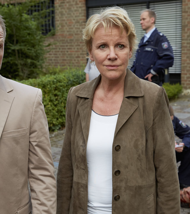 "Marie Brand und das ewige Wettrennen": Marie Brand (Mariele Millowitsch) und Jürgen Simmel (Hinnerk Schönemann) stehen in einer Einfahrt vor einem Einfamilienhaus. Hinter ihnen liegt das Opfer auf dem Boden. Weitere Beamten in Uniform sind zu sehen.