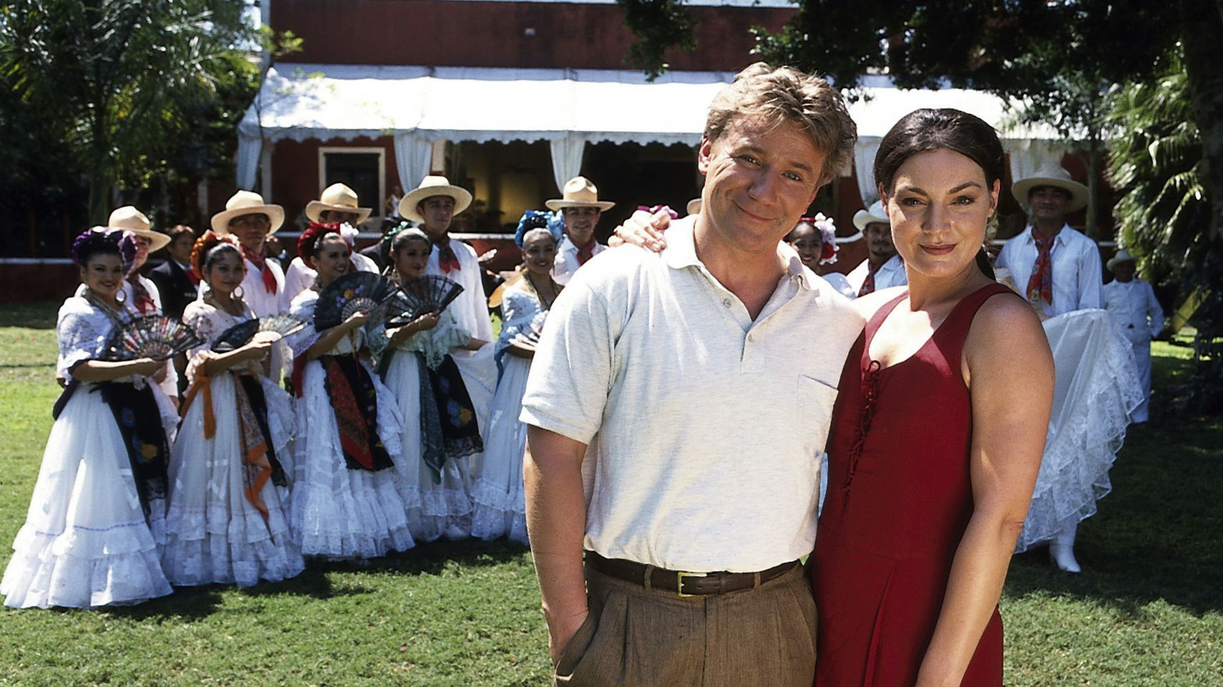 "Das Traumschiff - Mexiko": Dr. Andreas Brauer (Jörg Schüttauf) steht Arm in Arm mit Corinna (Marion Mitterhammer) auf einer Wiese. Hinter ihnen steht eine argentinische Folkloregruppe.