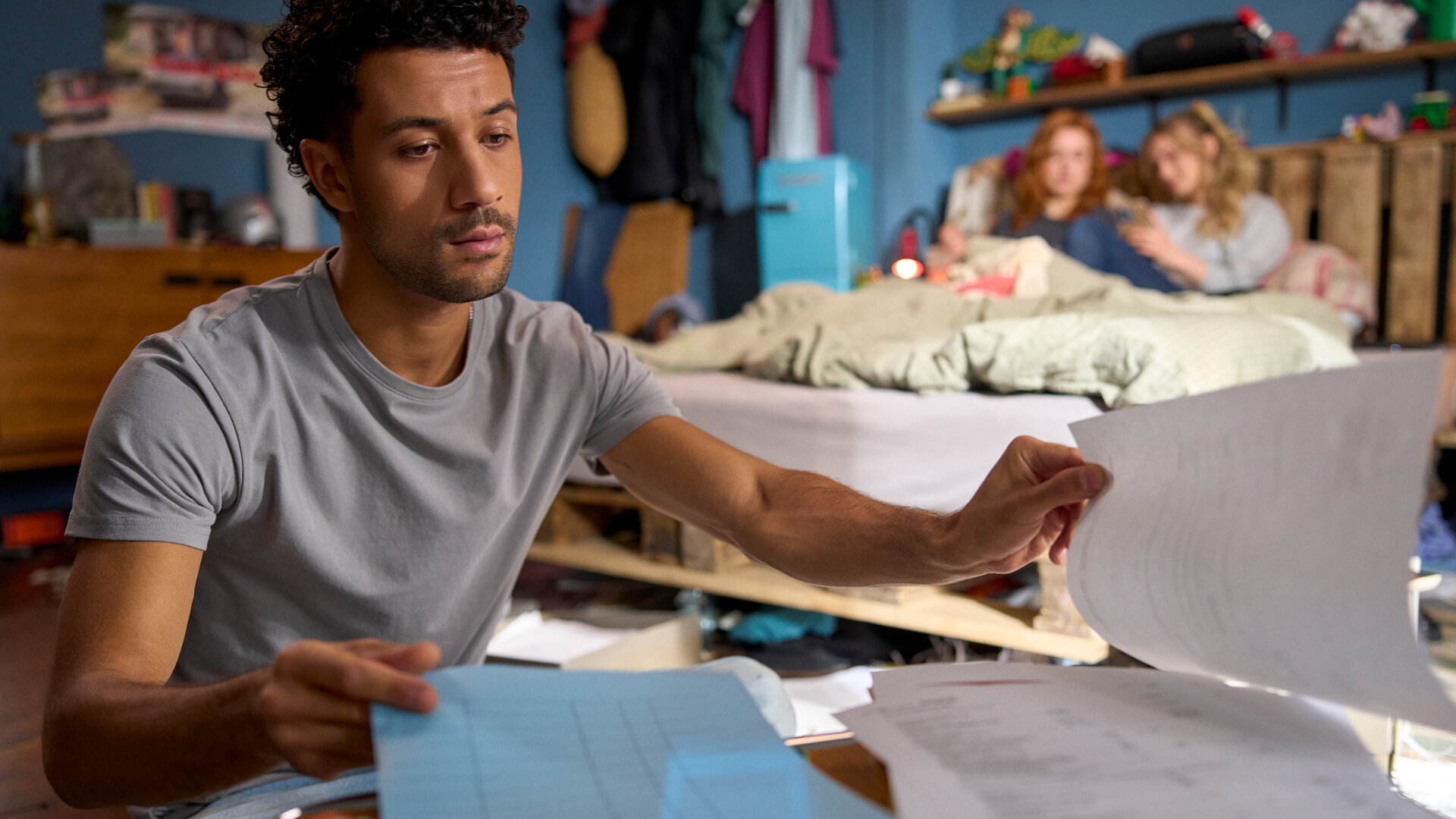 "Ich dich auch! - Das Gewitter": In einem Wohnraum: Yannik (Rojan Juan Barani) sitzt konzentriert vor einem Haufen Papiere. Caro (Meriel Hinsching) und Svenja (Leonie Wesselow) chillen auf dem Bett im Hintergrund.