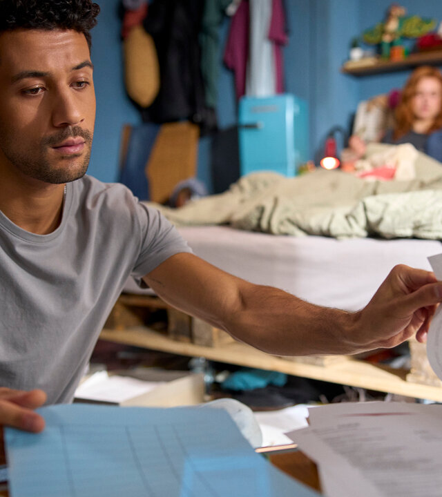 "Ich dich auch! - Das Gewitter": In einem Wohnraum: Yannik (Rojan Juan Barani) sitzt konzentriert vor einem Haufen Papiere. Caro (Meriel Hinsching) und Svenja (Leonie Wesselow) chillen auf dem Bett im Hintergrund.