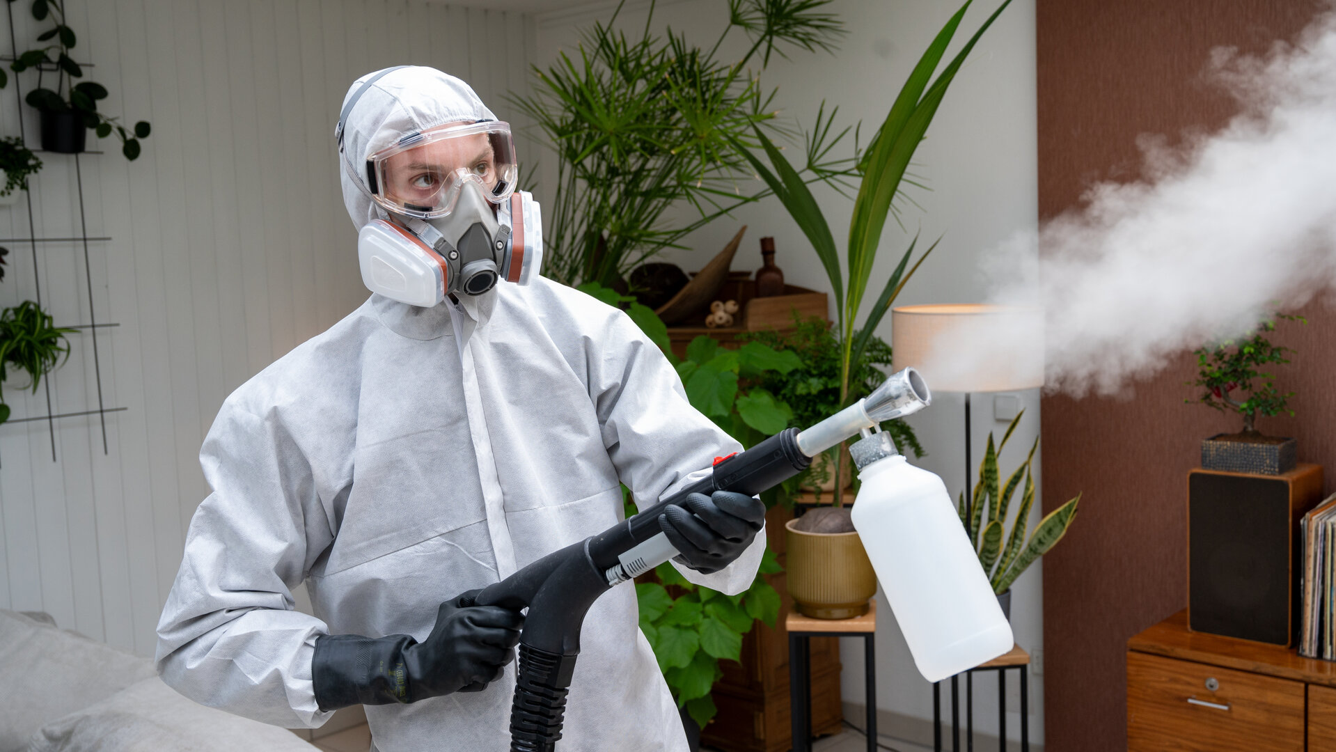 "SOKO Stuttgart - Der Saubermann": Der Tatortreiniger Benno Kruse (Kim Patrick Biele) steht in einem weißen Arbeitsoverall, einer Atemschutzmaske, Schutzbrille und schwarzen Handschuhen in einem Wohnraum und versprüht mit einem Profi-Sprühgerat Dunst in die Luft.