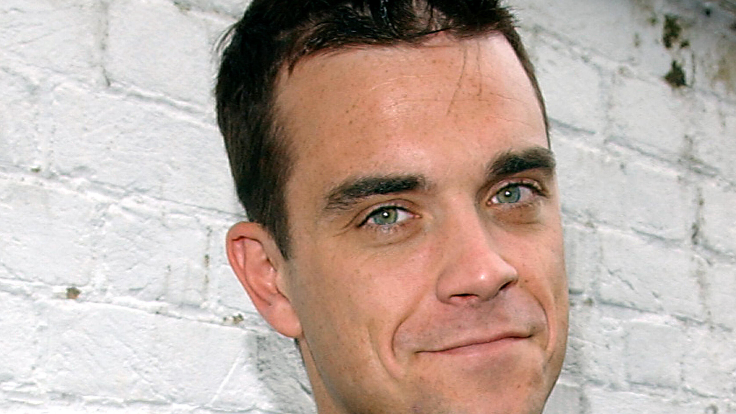 „The True Story of Robbie Williams": Robbie Williams steht vor einer Steinmauer und lächelt in die Kamera.