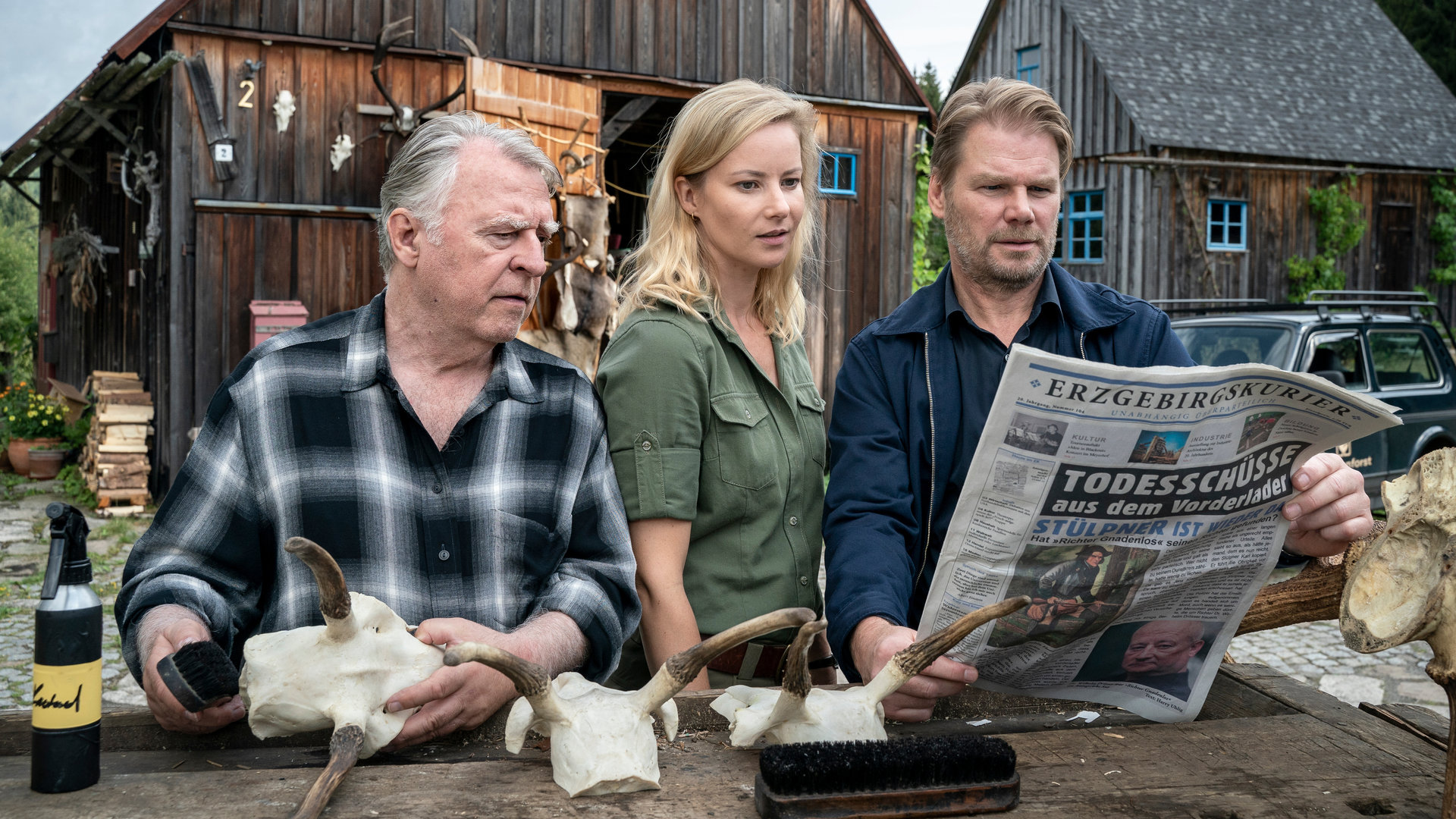 "Erzgebirgskrimi - Der letzte Bissen": Außenaufnahme. Georg Bergelt (Andreas Schmidt-Schaller), Saskia Bergelt (Teresa Weißbach) und Robert Winkler (Kai Scheve) stehen im Hof des Forstamtes an einem Tisch mit Geweihen und schauen in eine Zeitung, deren Schlagzeile lautet: "Todesschuss mit Vorderlader".