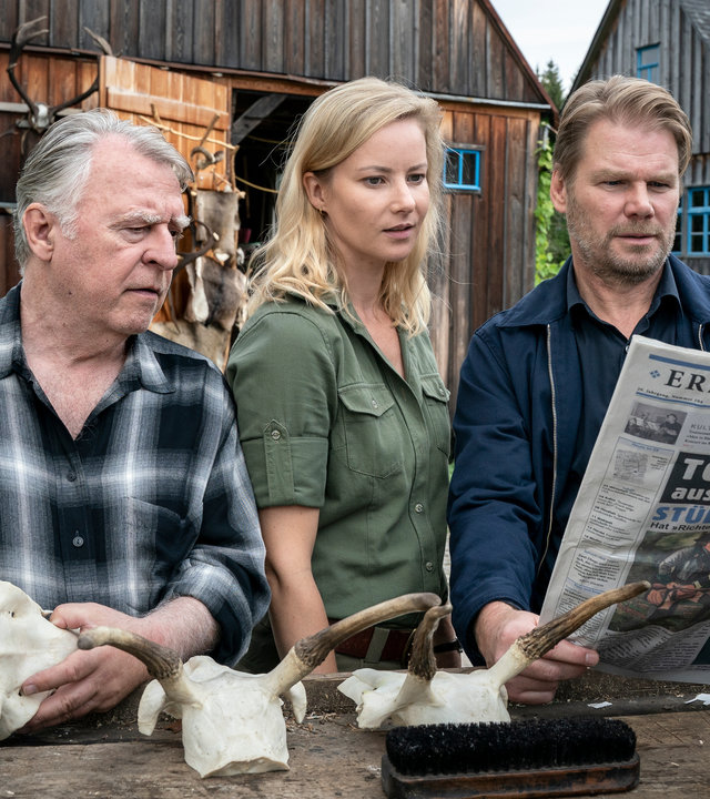 "Erzgebirgskrimi - Der letzte Bissen": Außenaufnahme. Georg Bergelt (Andreas Schmidt-Schaller), Saskia Bergelt (Teresa Weißbach) und Robert Winkler (Kai Scheve) stehen im Hof des Forstamtes an einem Tisch mit Geweihen und schauen in eine Zeitung, deren Schlagzeile lautet: "Todesschuss mit Vorderlader".