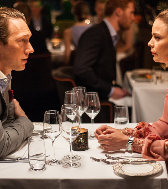 "Exit - Lügen": Adam Veile (Simon J. Berger) und Hermine Veile (Agnes Kittelsen) sitzen sich am gedeckten Tisch in einem Restaurant gegenüber. Beide blicken sich ernst an.