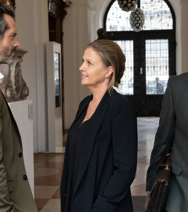 "Die Chefin - Söhne": Vera Lanz (Katharina Böhm) und Hannes de Boer (Martin Rapold) stehen sich in einem Flur gegenüber und lächeln sich an. Richter Dr. Jakob Murnau (Ernst Stötzner) schaut sie an und geht an ihnen vorbei.