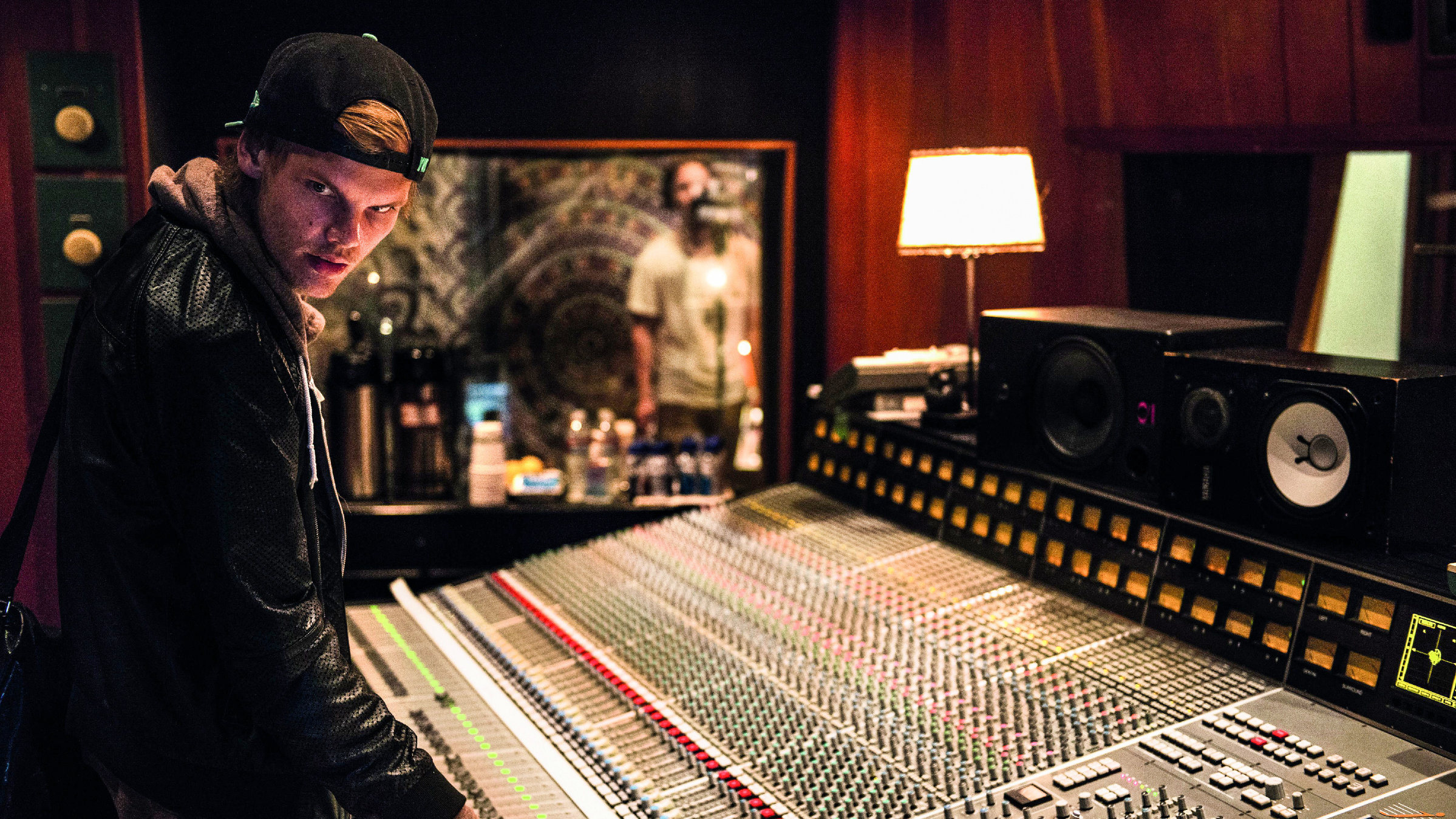"Avicii: Tribute Concert - In Loving Memory of Tim Bergling": Ein junger Mann steht an einem Mischpult in einem Tonstudio. Er trägt eine schwarze Lederjacke und ein Basecap. Er steht seitlich und hat den Kopf gedreht, so dass er in Richtung Kamera blickt.