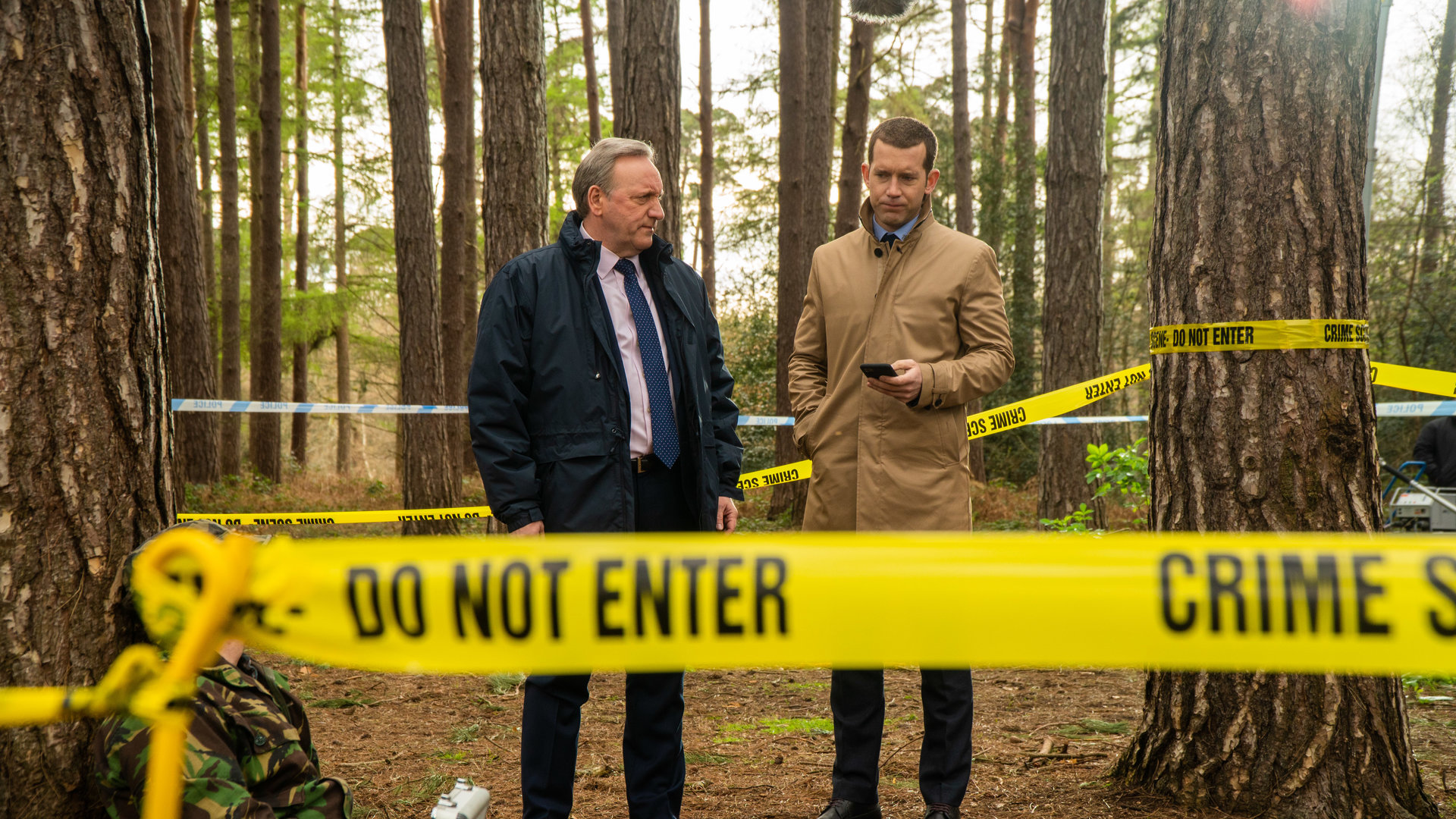 "Inspector Barnaby: Weltuntergang für einen Prepper": DCI John Barnaby (Neil Dudgeon) und sein Assistent DS Jamie Winter (Nick Hendrix) stehen in einem Waldstück umgeben von gelbem Polizeiabsperrband. Vor ihnen sitzt eine Person an einen Baum gelehnt.