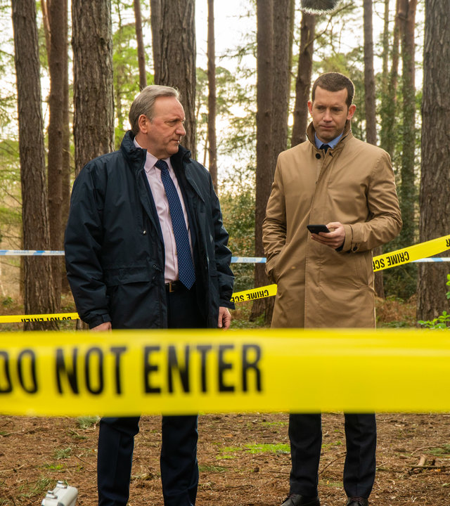 "Inspector Barnaby: Weltuntergang für einen Prepper": DCI John Barnaby (Neil Dudgeon) und sein Assistent DS Jamie Winter (Nick Hendrix) stehen in einem Waldstück umgeben von gelbem Polizeiabsperrband. Vor ihnen sitzt eine Person an einen Baum gelehnt.