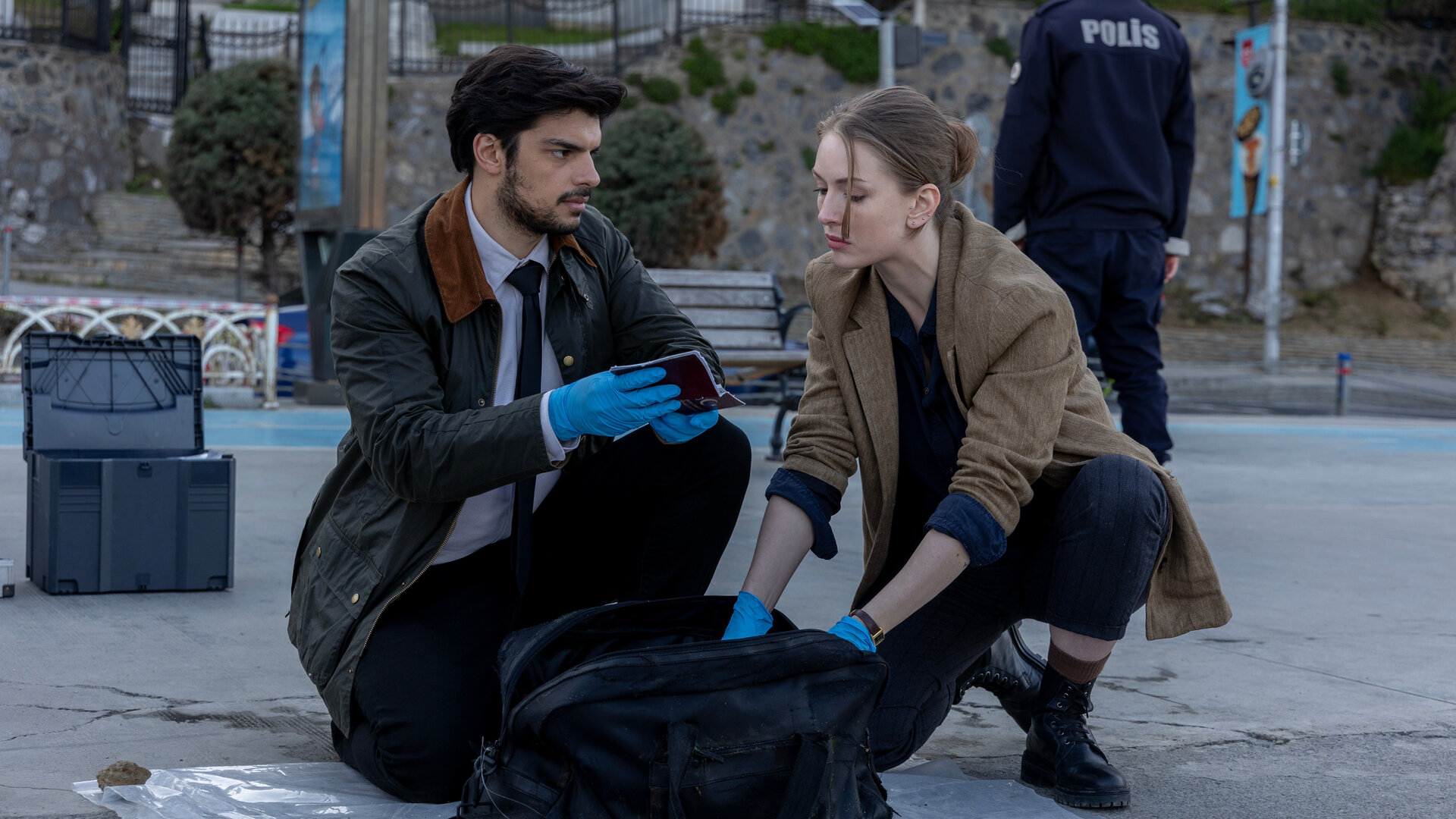 "Inspector Ikmen - Tod in Istanbul: Willkommen in Istanbul (2)": Tagsüber auf der Straße: Mehmet (Ethan Kai) und Ayse (Yasemin Kay Allen) hocken in ziviler Kleidung auf der Straße. Sie tragen blaue Plastikhandschuhe und durchsuchen eine schwarze Henkeltasche, die zwischen ihnen steht.