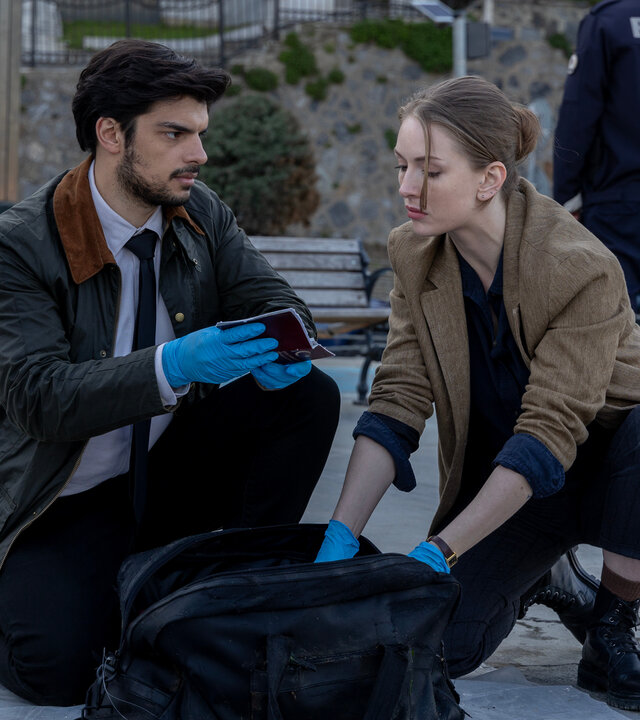 "Inspector Ikmen - Tod in Istanbul: Willkommen in Istanbul (2)": Tagsüber auf der Straße: Mehmet (Ethan Kai) und Ayse (Yasemin Kay Allen) hocken in ziviler Kleidung auf der Straße. Sie tragen blaue Plastikhandschuhe und durchsuchen eine schwarze Henkeltasche, die zwischen ihnen steht.