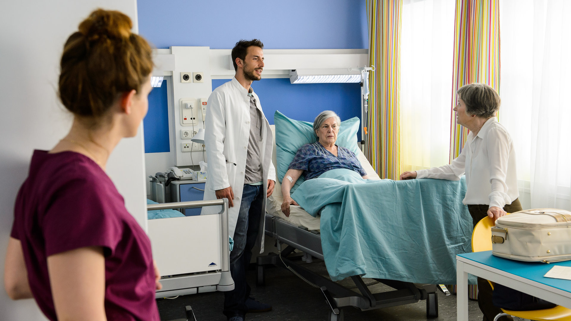 "Bettys Diagnose: Von Liebe und Leid": Hanna Winter (Marie Zielcke) steht an der Tür zum Krankenzimmer und beobachtet Dr. Frank Stern (Max Alberti), wie er mit seiner Oma Ilse Stern (Monika Lennartz) und deren Freundin Marlies Benz (Margot Gödrös) spricht.