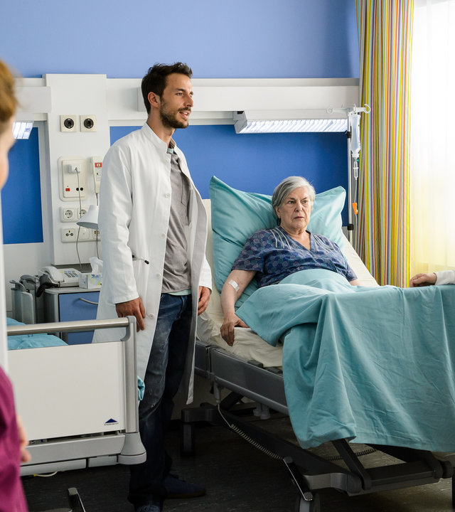 "Bettys Diagnose: Von Liebe und Leid": Hanna Winter (Marie Zielcke) steht an der Tür zum Krankenzimmer und beobachtet Dr. Frank Stern (Max Alberti), wie er mit seiner Oma Ilse Stern (Monika Lennartz) und deren Freundin Marlies Benz (Margot Gödrös) spricht.