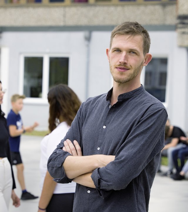 "37°: Kaltstart im Klassenzimmer - Quereinsteiger als Lehrer": Till (35) steht mit verschränkten Armen auf dem Schulhof. Um ihn herum sind Schüler und Schülerinnen in der Hofpause.