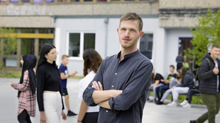 "37°: Kaltstart im Klassenzimmer - Quereinsteiger als Lehrer": Till (35) steht mit verschränkten Armen auf dem Schulhof. Um ihn herum sind Schüler und Schülerinnen in der Hofpause.