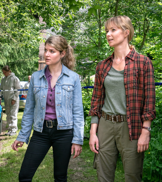 "Erzgebirgskrimi - Verhängnisvolle Recherche": Hauptkommissar Robert Winkler (Kai Scheve), Kommissarin Karina Szabo (Lara Mandoki), Försterin Saskia Bergelt (Teresa Weißbach) und Alt-Förster Georg Bergelt (Andreas Schmidt-Schaller) stehen in einer Reihe vor einer Leiche in einem baumbestandenen Garten und erörtern eine Frage.