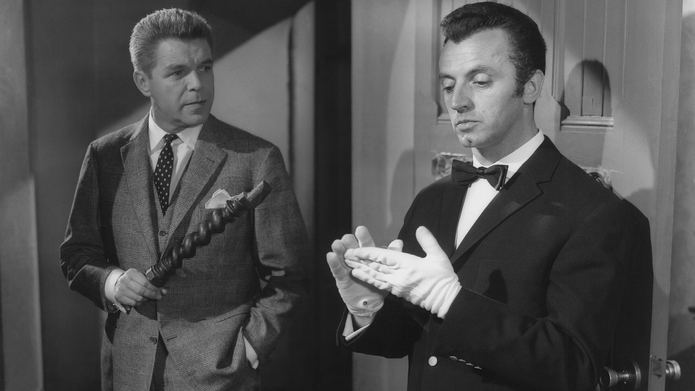 „Edgar Wallace: Das indische Tuch“: Der Anwalt Frank Tanner (Heinz Drache, links) hält eine Art hölzernes Möbelstück in der Hand, während er mit dem Butler Bonwit (Eddy Arendt) spricht, der sich gerade die weißen Handschuhe glattstreicht.