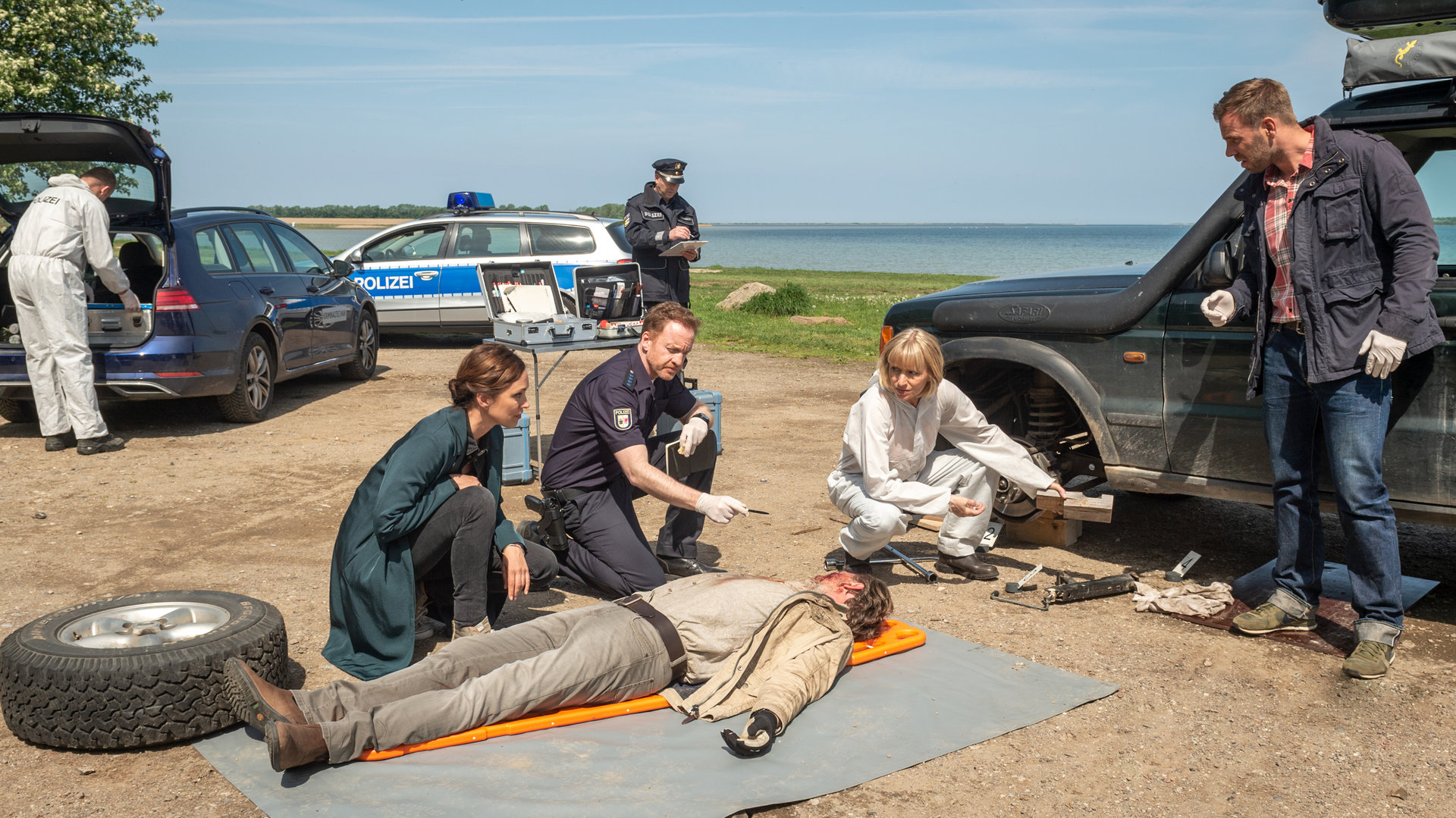 „SOKO Wismar – Lebe lieber unterirdisch“: Andreas Hidde (Marco Wittorf) liegt leblos auf einer Plane neben seinem Geländewagen. Der vordere Reifen des Wagens ist abmontiert und liegt zu seinen Füßen. Um den Toten herum hocken Karoline Joost (Nike Fuhrmann), Timmermann (Mathias Junge) und Roswitha Prinzler (Silke Matthias). Lars Pöhlmann (Dominic Boeer) steht ebenfalls dabei. Alle blicken zum Gesicht des Toten.