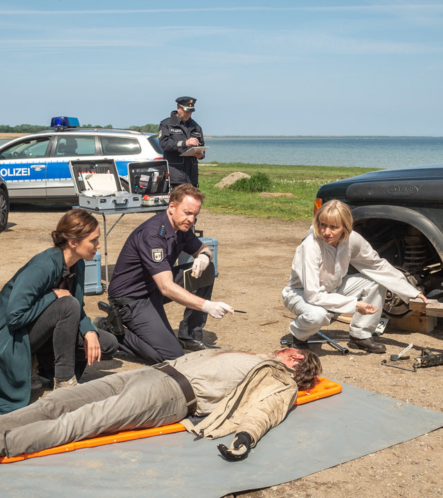 „SOKO Wismar – Lebe lieber unterirdisch“: Andreas Hidde (Marco Wittorf) liegt leblos auf einer Plane neben seinem Geländewagen. Der vordere Reifen des Wagens ist abmontiert und liegt zu seinen Füßen. Um den Toten herum hocken Karoline Joost (Nike Fuhrmann), Timmermann (Mathias Junge) und Roswitha Prinzler (Silke Matthias). Lars Pöhlmann (Dominic Boeer) steht ebenfalls dabei. Alle blicken zum Gesicht des Toten.