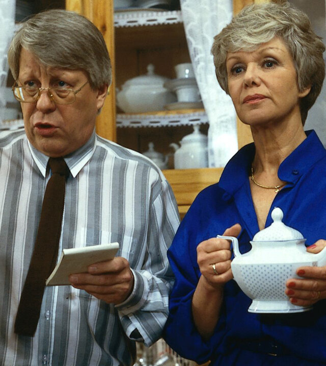 "Die Wicherts von nebenan: Käthes Küche": Eberhart (Stefan Orlac) und Hannelore (Maria Sebaldt) stehen in der Küche. Eberhard hält einen Notizblock und einen Stift in den Händen, Hannelore eine Teekanne.