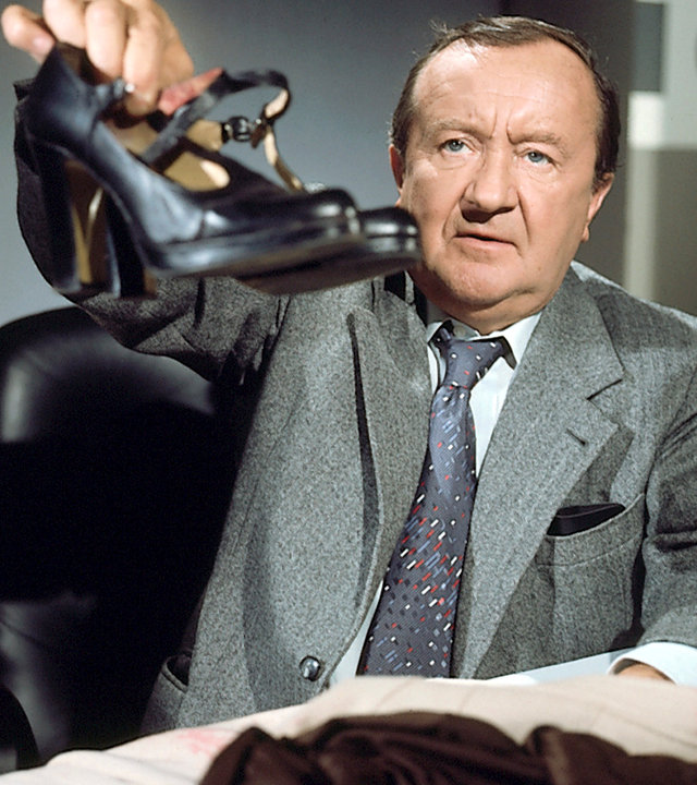 "Der Alte - Toccata und Fuge": Hauptkommissar Köster (Siegried Lowitz) sitzt an einem mit Kleider belegten Schreibtisch und einem Telefon, ganz rechts im Bild. Eine Lesebrille liegt auch noch auf dem Tisch. Er hält ein paar schwarze Damenschuhe in die Kamera.