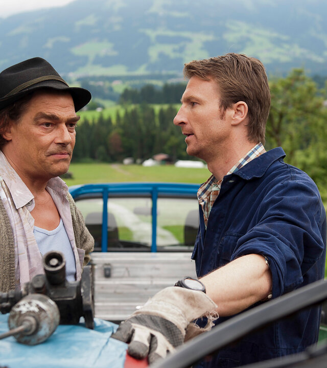 "Der Bergdoktor - Ausgeträumt": Arthur Distelmeier (Martin Feifel) und Gruber (Heiko Ruprecht) stehen auf einer Weide an einem Traktor und unterhalten sich. Im Hintergrund Berge.