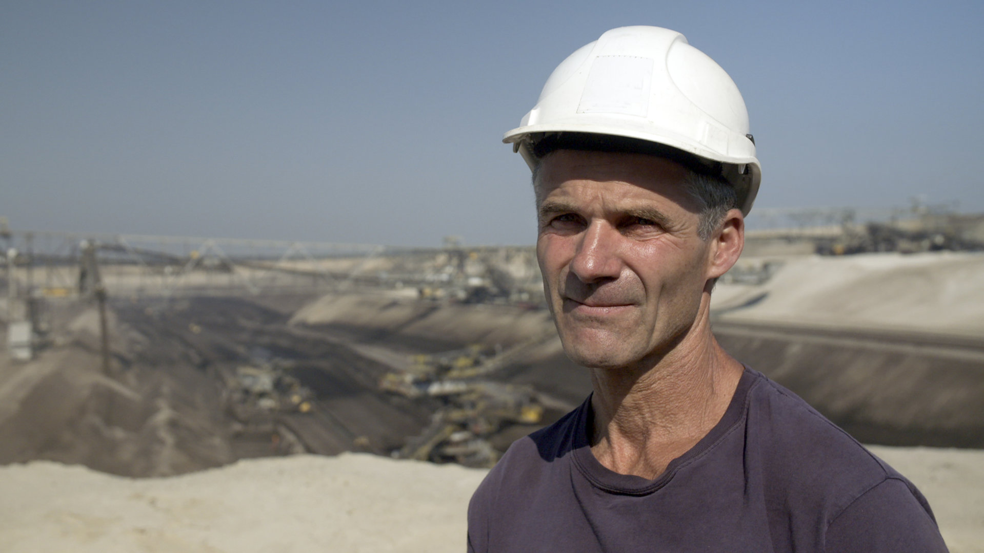 "37° Abgebaggert - Leben ohne Kohle": Michael steht an der Kante zu einem Tagebau eines Kohlewerks. Er trägt einen weißen Bauhelm und schaut in die Kamera.