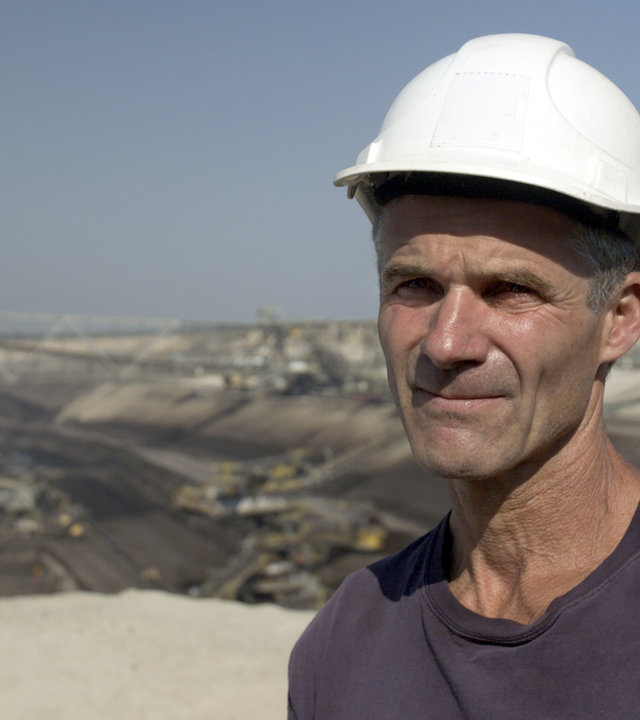 "37° Abgebaggert - Leben ohne Kohle": Michael steht an der Kante zu einem Tagebau eines Kohlewerks. Er trägt einen weißen Bauhelm und schaut in die Kamera.