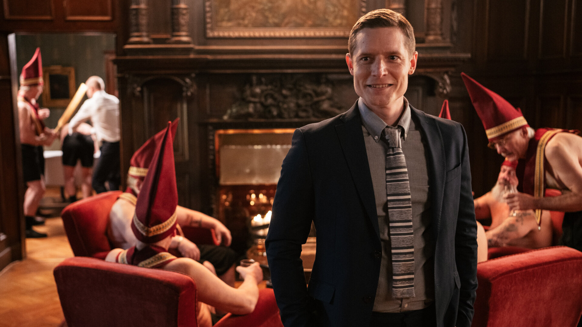 "Queen of Fucking Everything - Dünnes Eis": Mikko (Max Ovaska) steht in einem edel eingerichteten Raum und blickt in Richtung Kamera. Hinter ihm sind mehrere Männer mit nackten Oberkörpern zu sehen, die hohe rote Mützen und Schärpen mit Goldrand tragen. Mikko selbst trägt einen Anzug und Krawatte.