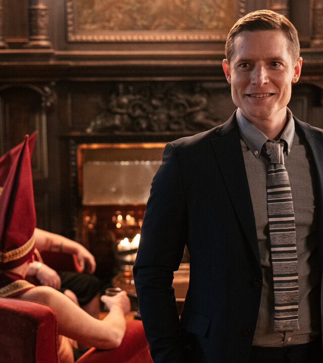 "Queen of Fucking Everything - Dünnes Eis": Mikko (Max Ovaska) steht in einem edel eingerichteten Raum und blickt in Richtung Kamera. Hinter ihm sind mehrere Männer mit nackten Oberkörpern zu sehen, die hohe rote Mützen und Schärpen mit Goldrand tragen. Mikko selbst trägt einen Anzug und Krawatte.