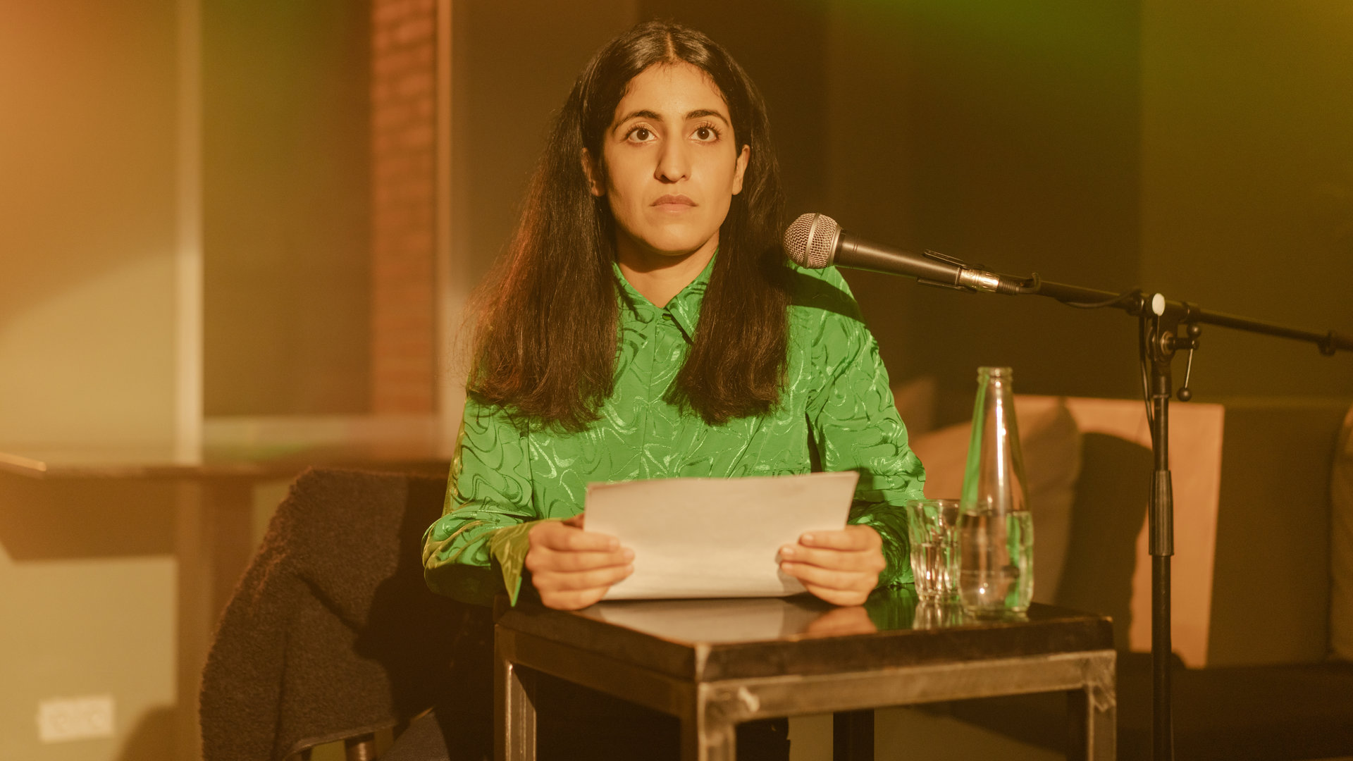 "Loving Her - Loving You": Hanna (Banafshe Hourmazdi) steht an einem Pult. Seitlich auf dem Pult steht eine Flasche Wasser und ein Glas. In ihren Händen hält sie einen Papierbogen. Ein Mikrofon ist auf sie gerichtet. Sie blickt überrascht nach vorne.