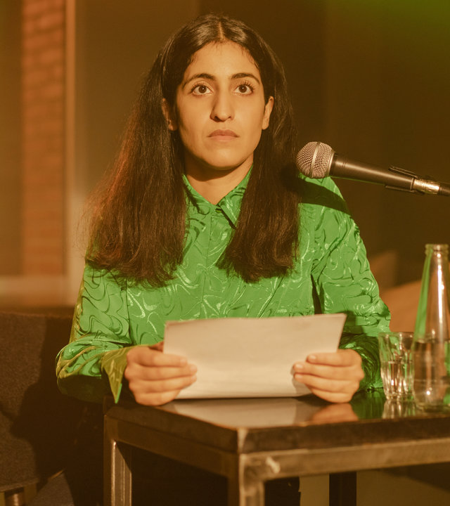 "Loving Her - Loving You": Hanna (Banafshe Hourmazdi) steht an einem Pult. Seitlich auf dem Pult steht eine Flasche Wasser und ein Glas. In ihren Händen hält sie einen Papierbogen. Ein Mikrofon ist auf sie gerichtet. Sie blickt überrascht nach vorne.