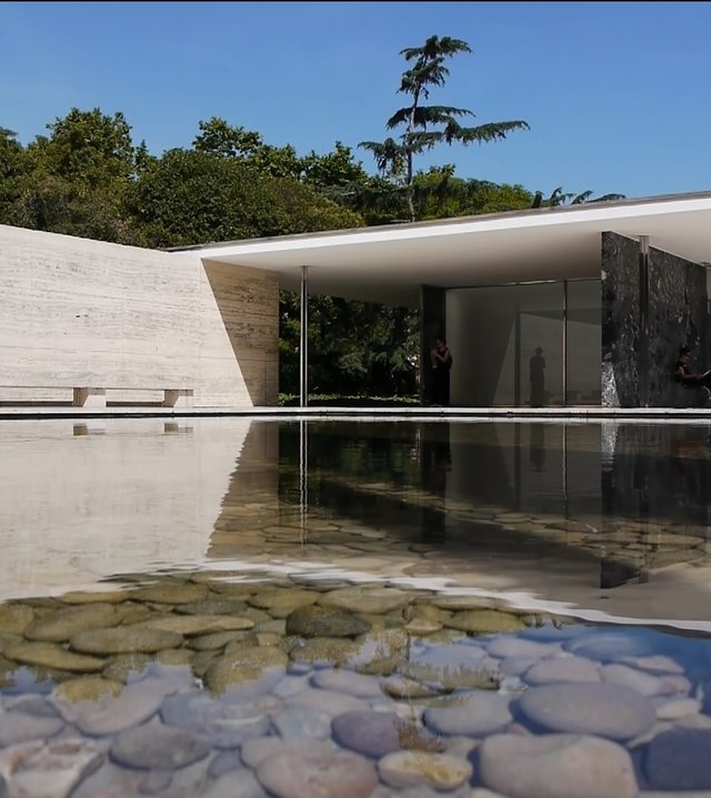 "Die Bauhaus-Revolution: Eleganter Flachdach-Bungalow, verglast, an einem Teich, im Wasser runde Steine. Dahinter Bäume.