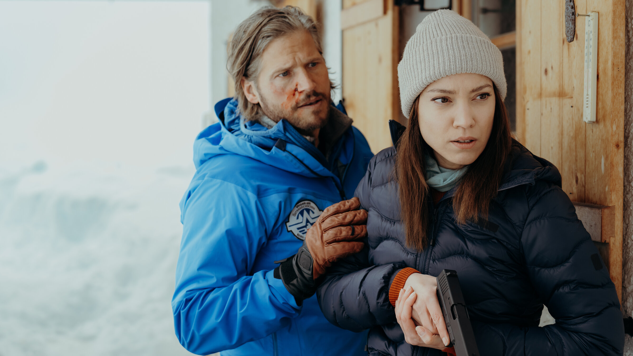 "Die Bergretter: Seelenfrieden": An einer Berghütte im Schnee: Mit der Dienstwaffe in der Hand steht Alex (Viktoria Ngotsé) angespannt vor der Tür. Mit blutender Gesichtswunde steht Markus Kofler (Sebastian Ströbel) hinter ihr und hat beruhigend seine Hand auf ihren Arm gelegt.