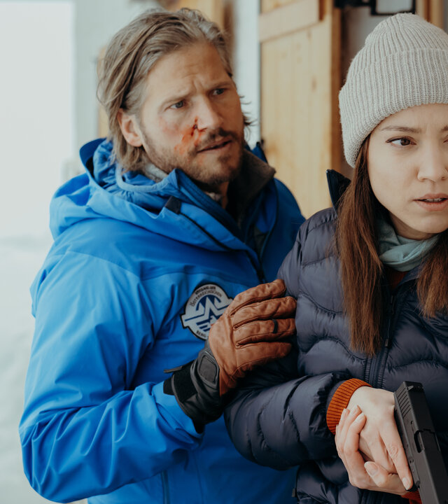"Die Bergretter: Seelenfrieden": An einer Berghütte im Schnee: Mit der Dienstwaffe in der Hand steht Alex (Viktoria Ngotsé) angespannt vor der Tür. Mit blutender Gesichtswunde steht Markus Kofler (Sebastian Ströbel) hinter ihr und hat beruhigend seine Hand auf ihren Arm gelegt.