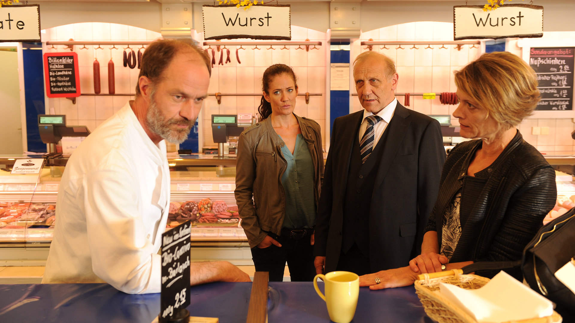 "SOKO Stuttgart - King of Vegan": Selma Kirsch (Yve Burbach) und Michael Kaiser (Karl Kranzkowski) stehen im Verkaufsraum der Metzgerei und reden mit Helena Meister (Karin Giegerich) und Willem Berg (Holger Daemgen), die nebeneinander an einem Stehtisch stehen.