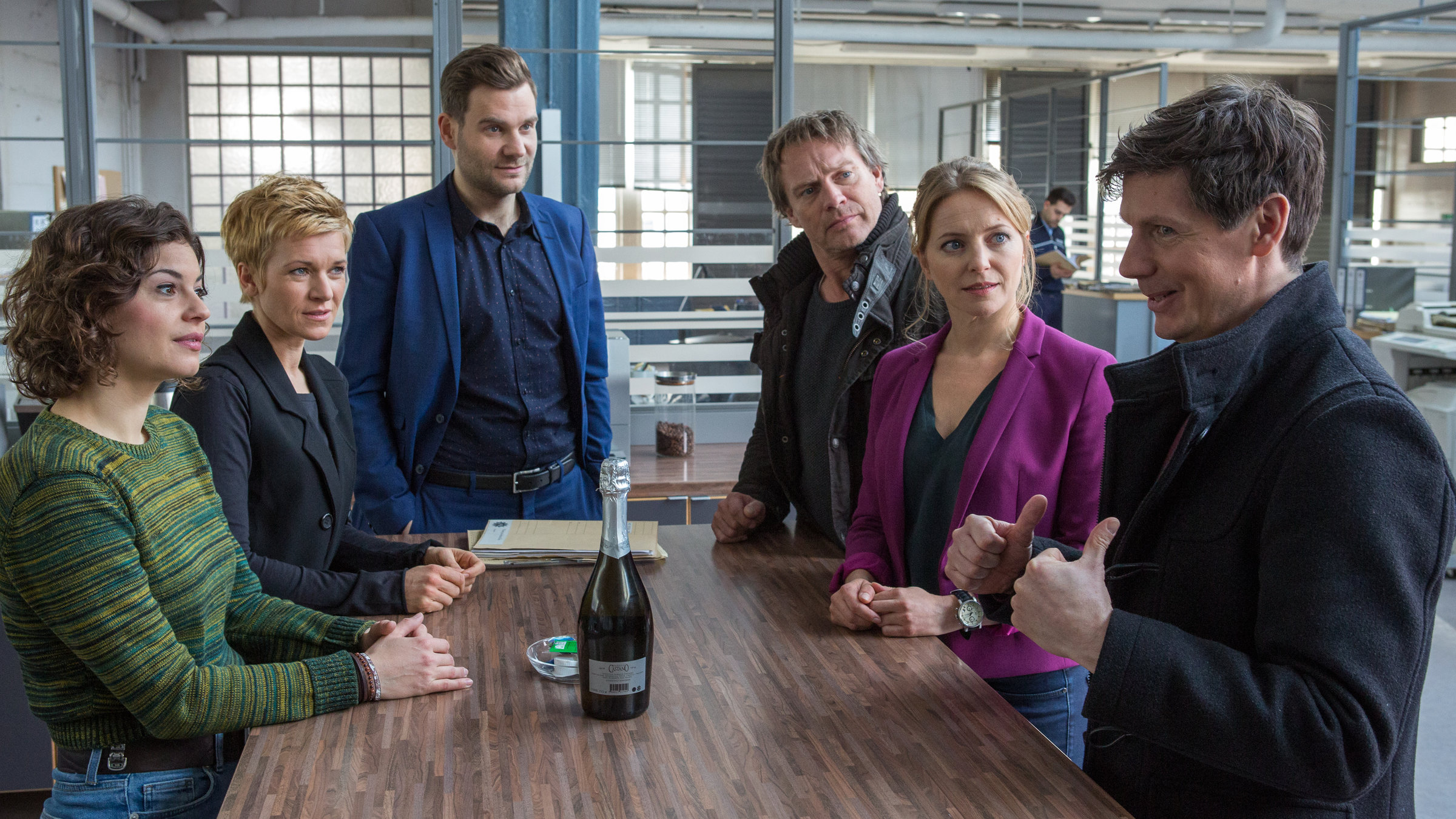 "SOKO Köln – Mülheimer Dämmerung“: Die Kommissare Jonas Fischer (Lukas Piloty), Lilly Funke (Tatjana Kästel), Vanessa Haas (Kerstin Landsmann), Matti Wagner (Pierre Besson) und Anna Maiwald (Diana Staehly) und Pathologe Dr. Philip Kraft (Thomas Clemens) stehen im Kommissariat versammelt um den Stehtisch. Auf dem Tisch steht eine geschlossene Flasche Sekt und es liegen Akten darauf. Alle schauen auf Dr. Philip Kraft, der gerade etwas erklärt und mit beiden Daumen nach oben zeigt.