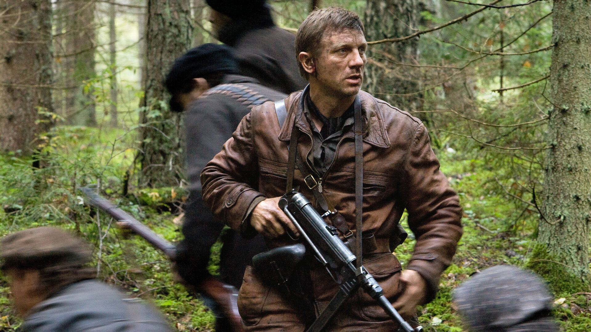 "Unbeugsam - Defiance": Tuvia (Daniel Craig) steht mit entschlossenem Blick in einem dichten Wald, eine Maschinenpistole im Anschlag, während um ihn herum Menschen (Komparsen) flüchten.