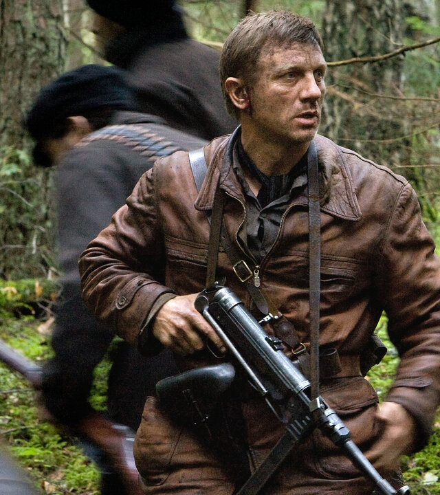 "Unbeugsam - Defiance": Tuvia (Daniel Craig) steht mit entschlossenem Blick in einem dichten Wald, eine Maschinenpistole im Anschlag, während um ihn herum Menschen (Komparsen) flüchten.