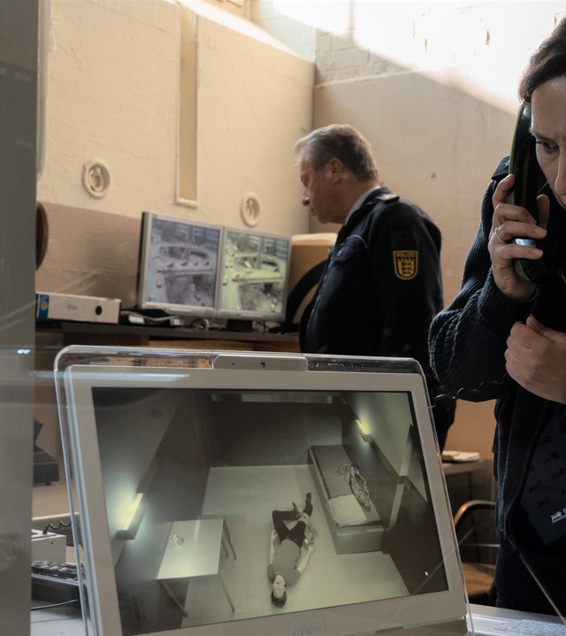 "SOKO Stuttgart – Goldregen": Streifenpolizistin Sibylle Beyer (Bärbel Stolz) steht in der Wache des Präsidiums und starrt geschockt auf den Bildschirm, auf dem das Bild der Überwachungskamera aus der Zelle zu sehen ist. Man sieht eine Person, die in der Zelle auf dem Boden liegt. Bille hat bereits den Telefonhörer am Ohr und meldet den Vorfall. Hinter ihr ist ein weiterer Beamter bei der Arbeit zu sehen.