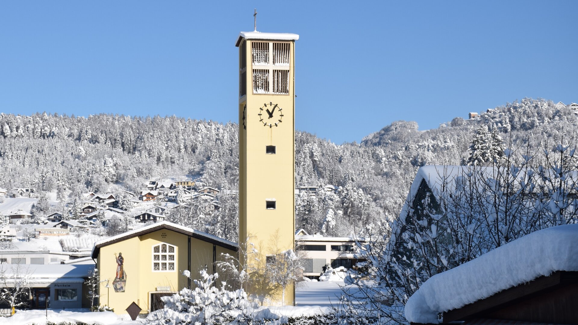 "Katholischer Gottesdienst - Was bleibt von Weihnachten?": Außenansicht der Pfarrkirche in Muntlix, Vorarlberg, in verschneiter Umgebung mit strahlend blauem Himmel.