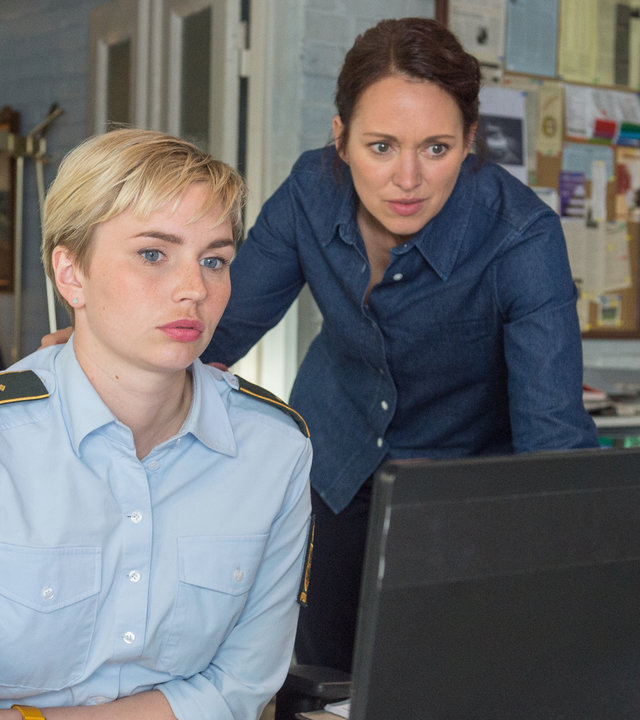 „SOKO Wismar – Scheiden tut weh“: Im Büro der Kripo sitzt Stine Bergendahl (Sidsel Hindhede) hinter ihrem Computer. Kommissarin Karoline Joost (Nike Fuhrmann) steht neben ihr, den Arm auf den Tisch gestützt. Beide schauen konzentriert auf den Bildschirm. Hinter ihnen stehen Regale mit Akten.