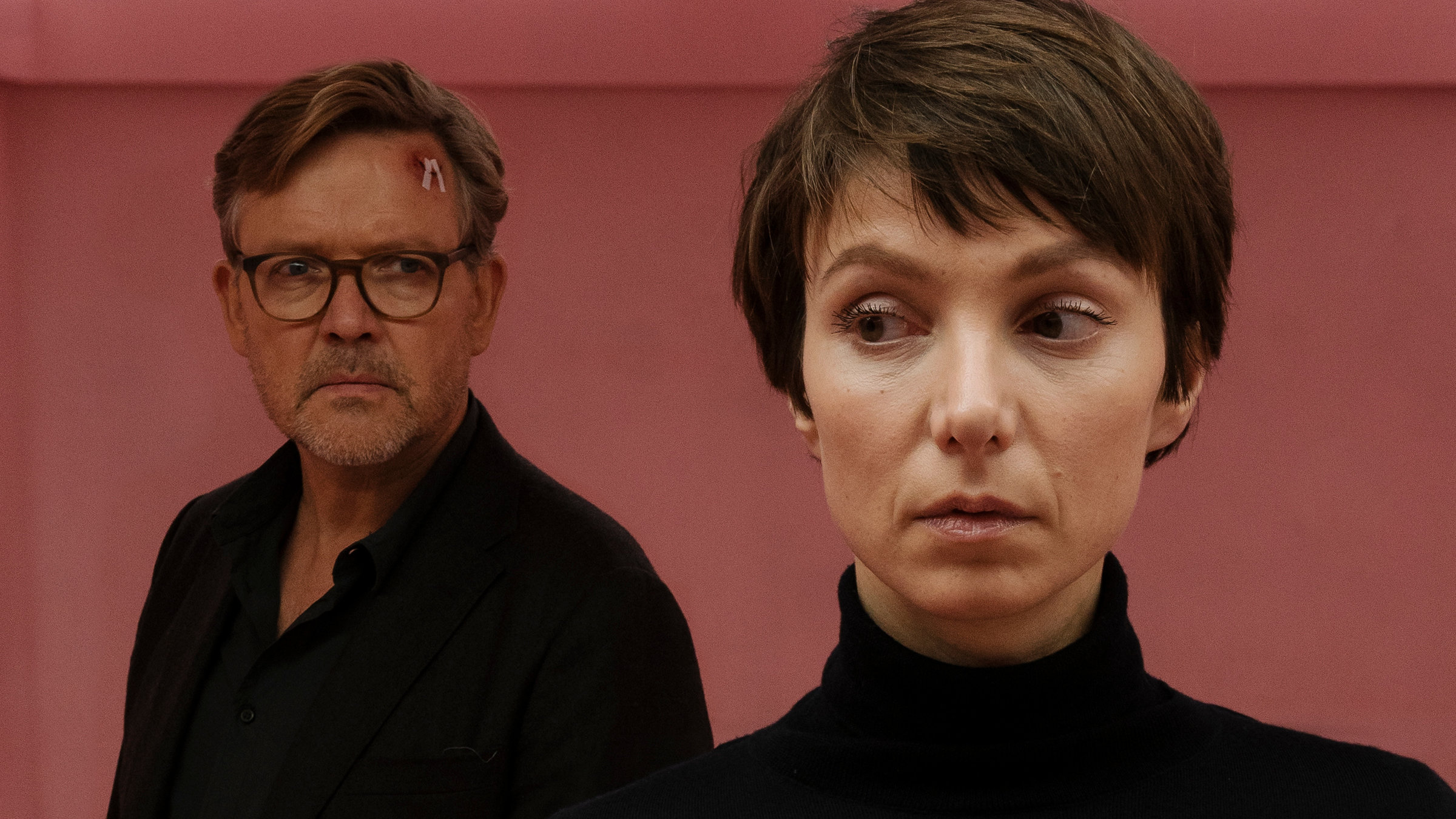 „Im Schatten der Angst“: Carsten Spanger (Justus von Dohnányi) steht mit einer Wunde am Kopf im Hintergrund vor einer rosa Wand und schaut auf Karla Eckhardt (Julia Koschitz), die den Kopf gerade, den Blick aber seitwärts gerichtet hat.