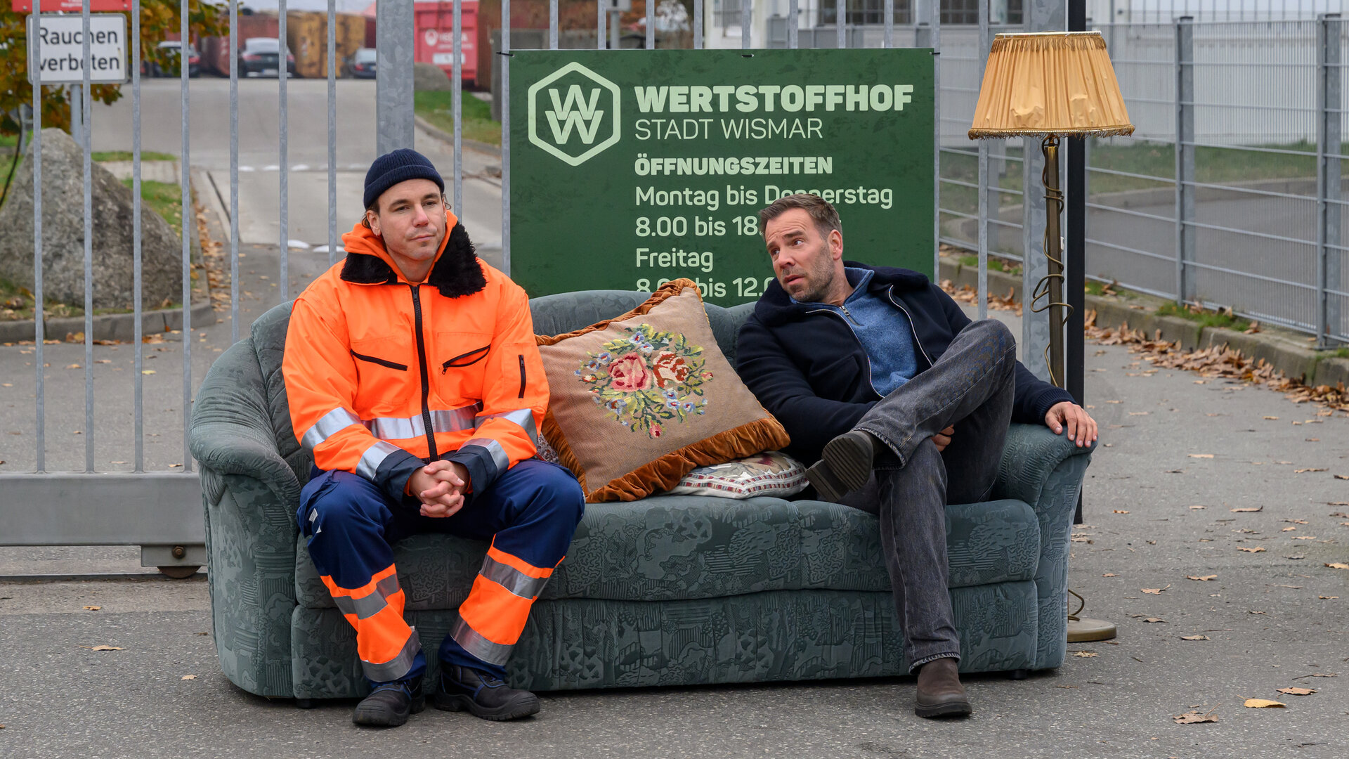 „SOKO Wismar – Schnelles Geld“: Vor dem Eingangstor des Wertstoffhofs sitzen Thore Stuck (Timo Weisschnur) und Lars Pöhlmann (Dominic Boeer) auf einem alten blauen Sofa, dass hier illegal abgeladen wurde. Thore trägt orange-blaue Arbeitskleidung. Neben Lars steht eine Stehlampe. Hinter ihnen hängt das Firmenschild am Tor.