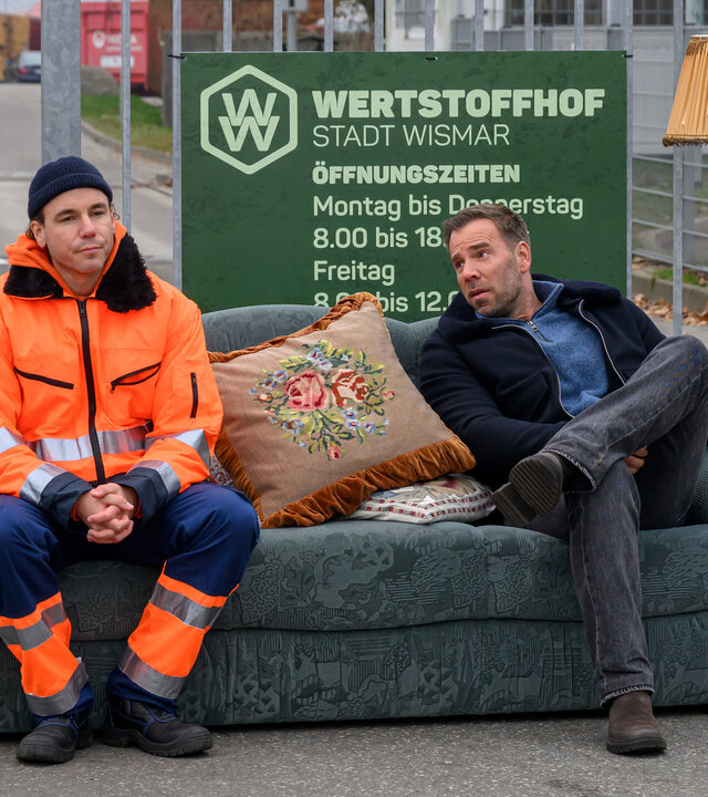 „SOKO Wismar – Schnelles Geld“: Vor dem Eingangstor des Wertstoffhofs sitzen Thore Stuck (Timo Weisschnur) und Lars Pöhlmann (Dominic Boeer) auf einem alten blauen Sofa, dass hier illegal abgeladen wurde. Thore trägt orange-blaue Arbeitskleidung. Neben Lars steht eine Stehlampe. Hinter ihnen hängt das Firmenschild am Tor.