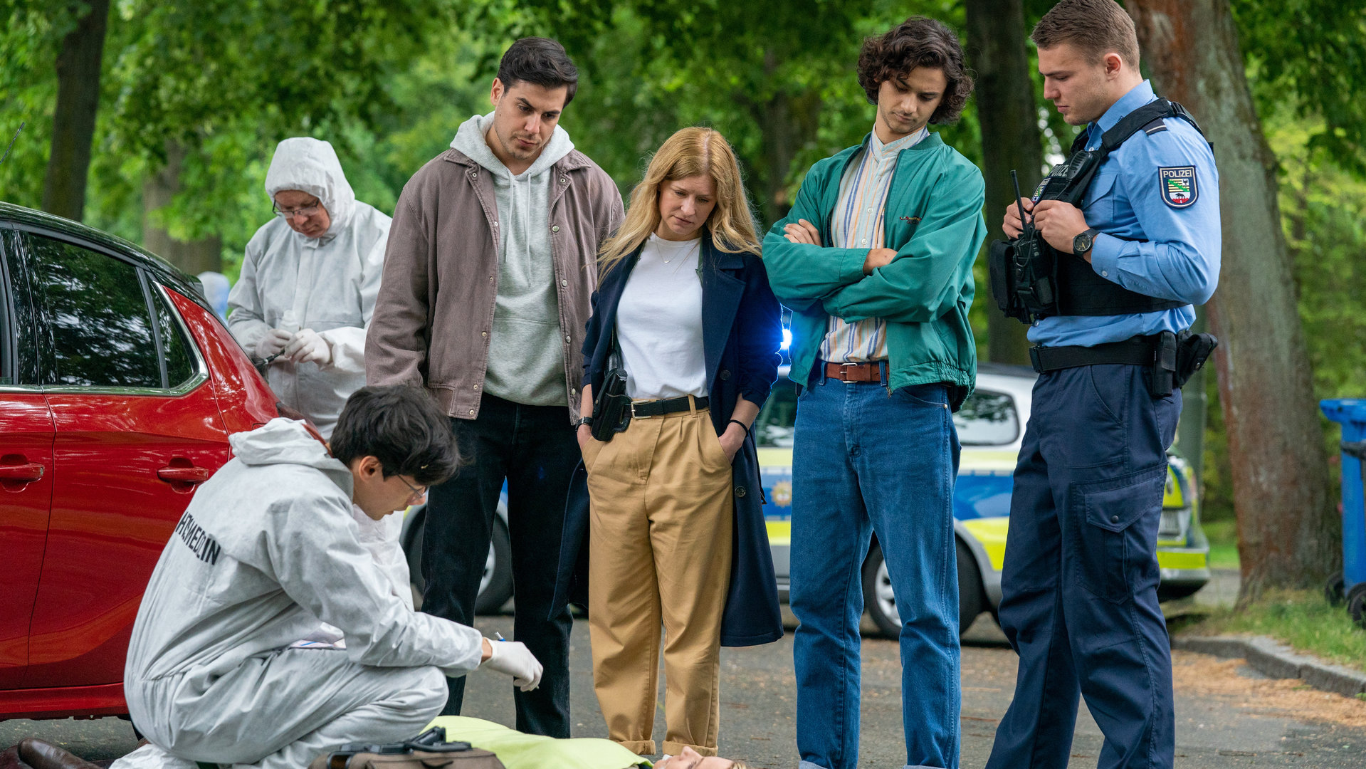 "Blutige Anfänger - Wut": Umut (Uğur Ekeroğlu), Koko (Birte Hanusrichter), Bruno (Martin Peñaloza Cecconi) und Phillip (Erik Cordes) stehen nebeneinander auf einer Straße. Alle blicken auf den Boden. Dort liegt Michelle Listing (Janine Theisen). Sie ist tot. Neben ihr in der Hocke sitzt Leon Landsberg (Florian Clyde). Er trägt einen weißen Overall der Spurensicherung und Handschuhe.