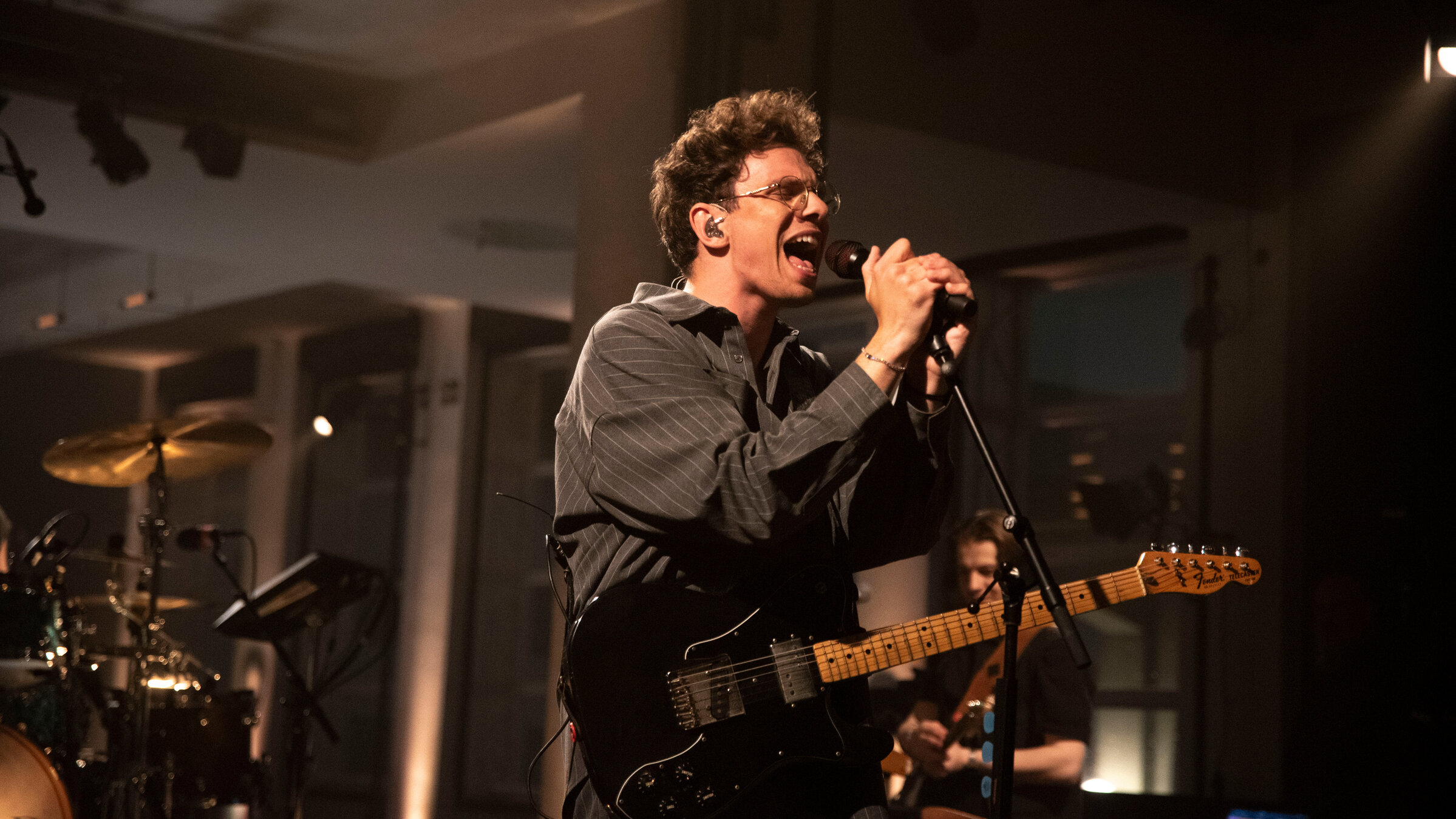 "Pop Around@Bauhaus: Kamrad": Der Sänger Kamrad steht auf der Bühne. Er hat eine E-Gitarre umhängen. Mit beiden Händen umfasst er das Mikrofon auf einem Mikrofonständer vor sich und singt. Bekleidet ist er mit einem braunen Hemd mit schmalen weißen Streifen. Im Hintergrund sieht man einen weiteren Musiker mit einer E-Gitarre und ein Schlagzeug.