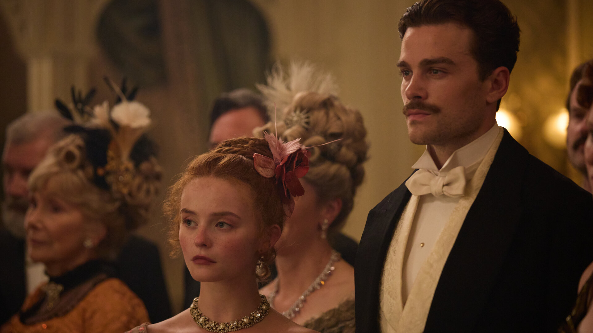 "The Forsytes - Familie verpflichtet: Wendepunkte": Auf einem Ball: Irene (Millie Gibson) und Soames (Joshua Orpin) stehen in eleganter Abendrobe nebeneinander und schauen mit ernstem Gesichtsausdruck seitlich nach vorne. Im Hintergrund stehen andere Personen (Komparsen) in Abendrobe.
