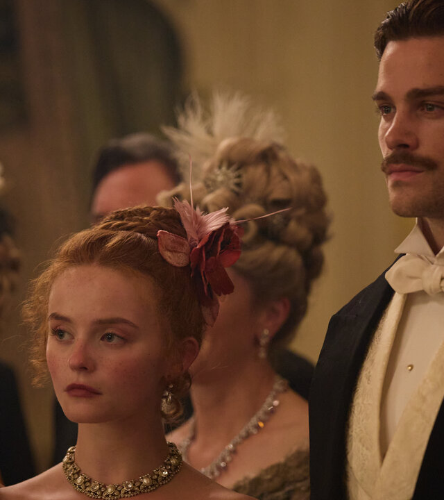 "The Forsytes - Familie verpflichtet: Wendepunkte": Auf einem Ball: Irene (Millie Gibson) und Soames (Joshua Orpin) stehen in eleganter Abendrobe nebeneinander und schauen mit ernstem Gesichtsausdruck seitlich nach vorne. Im Hintergrund stehen andere Personen (Komparsen) in Abendrobe.